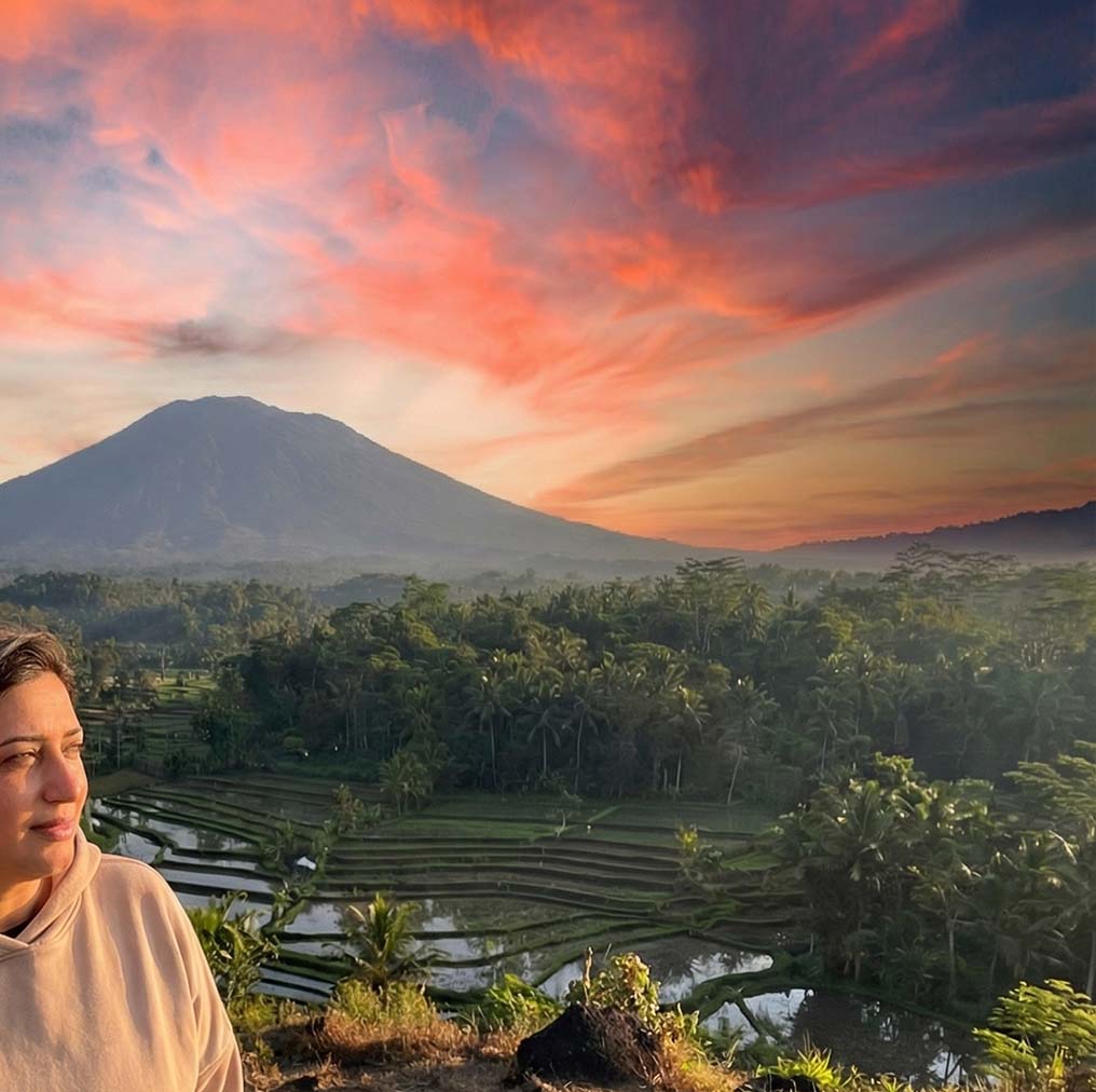 Person standing in front of a scenic view with rice terraces and a mountain under a colorful sky.