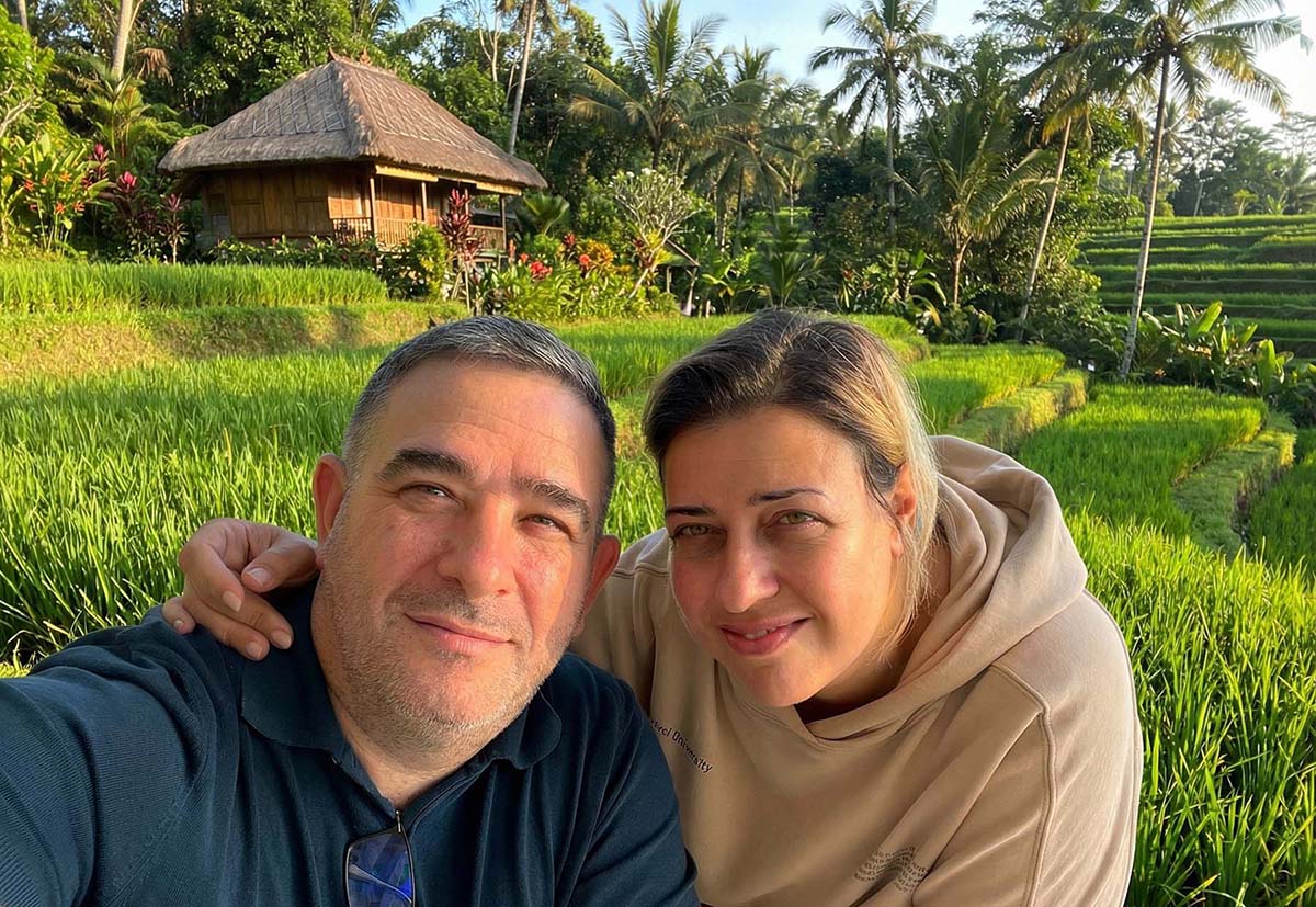 Two people taking a selfie in front of a rice paddy with a hut and trees in the background.